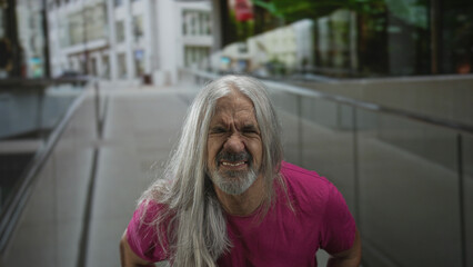 Man with long gray hair grimacing and showing teeth on an urban street near a glass building; defiance strength.