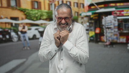 Man clasping hands to chest, smiling with red glasses and white shirt on street with scooters and...