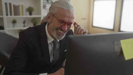 Man in suit adjusting glasses at office desk with computer monitor, smiling and leaning forward; confidence wisdom.