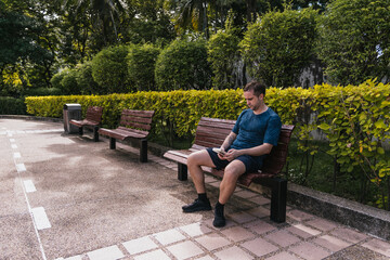 Fototapeta premium Man relaxing on a park bench holding a book. Green hedges and trees provide a natural outdoor setting