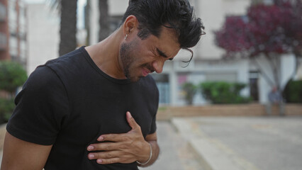 Young hispanic man clutch chest for heartattack on city street beside palm tree and building...