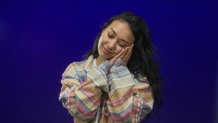 Young hispanic woman with hands to cheek for sleep pose in studio against deep cobalt blue purple wall wearing patterned jacket and hoop earrings; serenity.