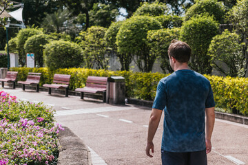 Man walking alone along a sunlit paved park path past wooden benches and lush green bushes,...
