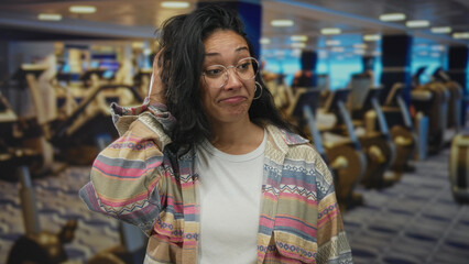Woman showing face and glasses smiling and shrugging shoulder in gym building, casual shirt and hoop earring; amusement.