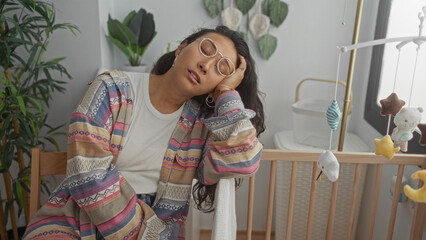 Woman resting head on hand beside wooden baby crib in building nursery room, wearing glasses and patterned cardigan; fatigue motherhood rest.