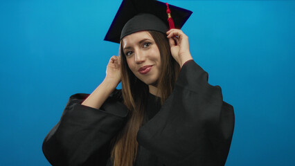 Young hispanic woman in graduation gown adjusts mortarboard tassel with her hands in studio;...