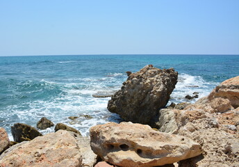 Vibrant summer view of the sea breaking against a rocky, sun-drenched shore. Natural stone texture in the foreground with clear blue water and sky. Vacation and travel concept.