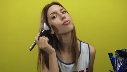 Young cheerleader in uniform applies makeup brush to cheek against yellow studio wall; confidence...