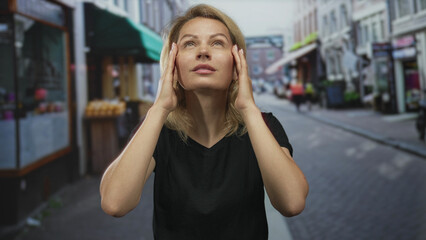 Young blonde woman pressing hands to forehead on busy city street with storefronts and pedestrians blurred behind her; stress.