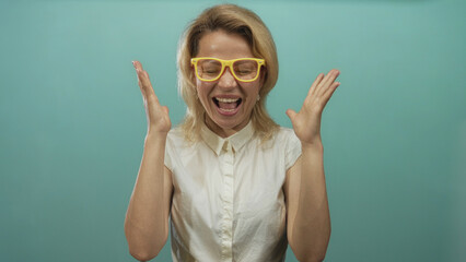 Blonde woman wearing yellow glasses clenches fists in teal studio; excitement celebration joy confidence.