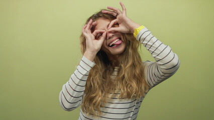Woman framing eyes with hands while peeking in studio as blonde young performer gives a smile and tongue expression against a green striped backdrop.
