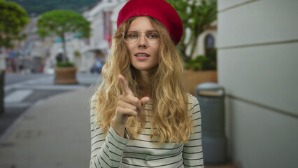 Fototapeta premium Scene captures woman hand shushing street with a blonde young finger pressing lips beneath red beret and glasses to emphasize silence.