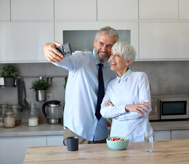 Portrait of a mid aged mature woman and man  gettyng ready for work using a smartphone and making a selfie photo  and eating breakfast oatmeal in a bowl standing in kitchen home. Overworked, busy, str