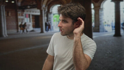 Fototapeta premium Man cups ear with hand to listen under arched city street arcade, smiling and leaning forward; curiosity attentiveness.