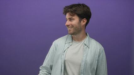 Obraz premium Man points thumb to side in a purple studio wearing light denim shirt and white tee, smiling while looking off camera; friendly casual.