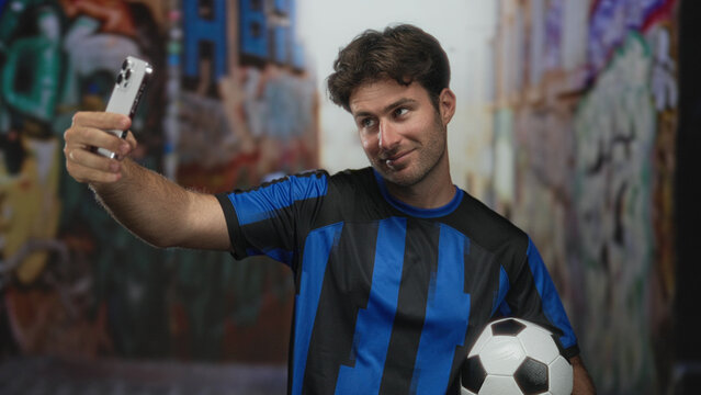 Young man holding smartphone and soccer ball while posing for a selfie in a graffiti street alley; team pride.