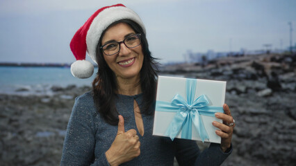 Obraz premium Middle age hispanic woman wearing santa hat and holding gift box with ribbon while giving thumb up on rocky seaside beach; holiday joy.
