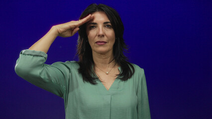 Fototapeta premium Woman wearing a pearl necklace and green blouse saluting with hand to forehead in studio with purple backdrop; duty.
