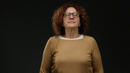 Woman practicing meditation with eyes closed, brunette hispanic middle-aged woman in orange sweater against isolated black wall background, representing calm and mindfulness.