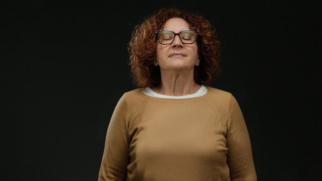 Woman practicing meditation with eyes closed, brunette hispanic middle-aged woman in orange sweater against isolated black wall background, representing calm and mindfulness.