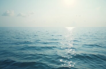 Vast calm ocean surface with gentle ripples and sun reflection. Clear blue water stretches to a hazy horizon under a pale sky with sparse clouds.