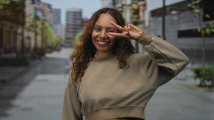 Woman showing peace sign with hand and smiling face on a lively city street with buildings; carefree joy.
