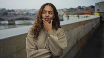 Fototapeta premium Woman biting fingernails on street beside old town bridge with hand to mouth gesture; anxiety uncertainty doubt.