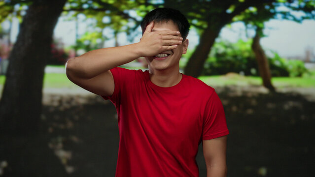 Young man in red shirt covers his eyes with hand while standing in a sunny park surrounded by lush green trees, creating a playful outdoor atmosphere. - Powered by Adobe