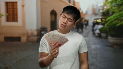 Young man in white shirt holds taiwanese banknotes on a bustling city street, reflecting on finances outdoors in urban taiwan.