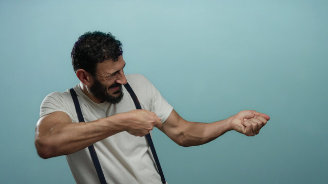 Hispanic man with beard pretending to pull rope against plain blue background, showcasing humorous action and expressive muscle tension.