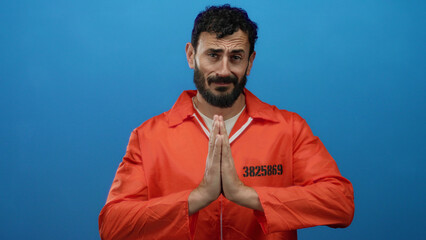 Hispanic man with beard wearing orange prison jumpsuit poses against a blue background in a meditative prayer position illustrating calm and introspection.