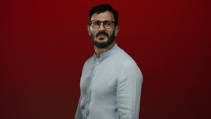 Hispanic man with beard and glasses stands against a bold red background in a casual shirt, exuding confidence and style.