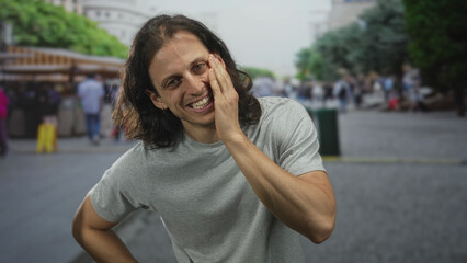Fototapeta premium Young hispanic man with hand on cheek smiling in busy street market with souvenir stalls and crowds in city outdoors; carefree joy.
