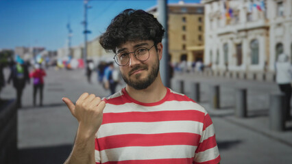 Young hispanic man with beard wearing glasses and striped shirt points thumb aside outdoors on a sunny day urban background showing casual attitude and style.