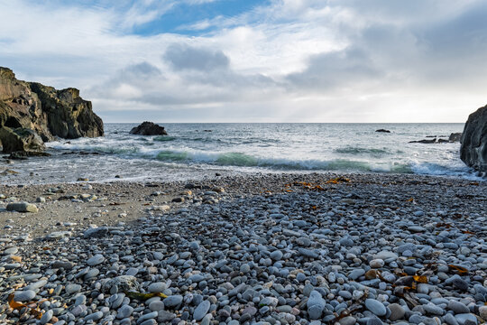 Pebble beach meets the sea at the foot of dramatic cliffs. The sky is cloudy.