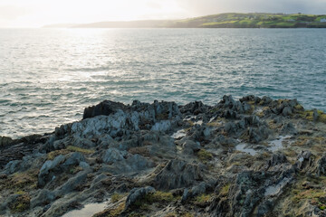 Jagged rocks meet the ocean in West Cork, Ireland. The sunlight glistens on the water, and green fields are visible in the distance on the horizon.