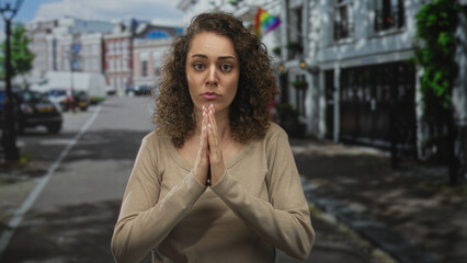 Fototapeta premium Young hispanic woman with hands pressed together praying on a city street in front of townhouses and storefronts; pleading hope.