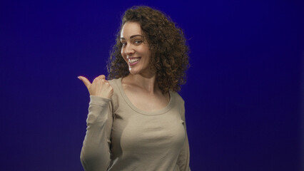 Woman wearing beige long sleeve top smiling and giving thumbs up while looking sideways in studio...
