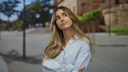 Fototapeta premium Young blonde woman quietly crosses arms in plaza by stone wall; rejection, doubt, skepticism, defiance.