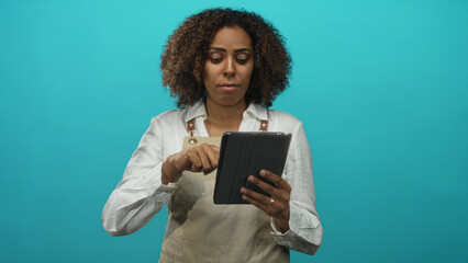Young african american woman baker wearing apron taps black tablet with right finger in teal studio; focus business planning.