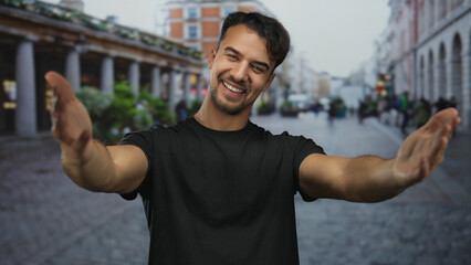 Fototapeta premium Young hispanic man smiling confidently on a bustling city street, offering a warm embrace amid urban vibrancy, showcasing a joyful outdoor scene.