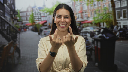 Fototapeta premium Woman smiling and beckoning with fingers on a cobblestone street in the city, wearing tied shirt and rings; joy connection.