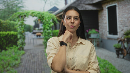 Woman points finger to camera at building entrance wearing beige shirt and smartwatch, hand on chin in thinking pose; thoughtful reflection.