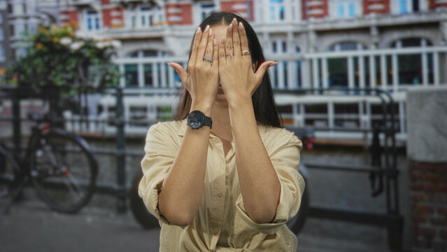 Woman with hands covering face and sticking tongue out on street in amsterdam canal district; playful spontaneous gesture.