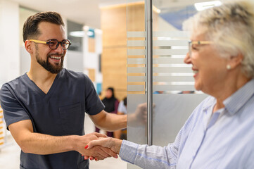 Male ophthalmologist opening glass door and greeting smiling senior woman with handshake at hospital entrance