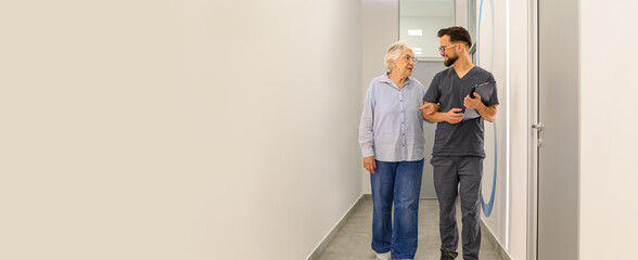 Senior woman walking arm in arm with male doctor in hospital hallway during medical consultation at...