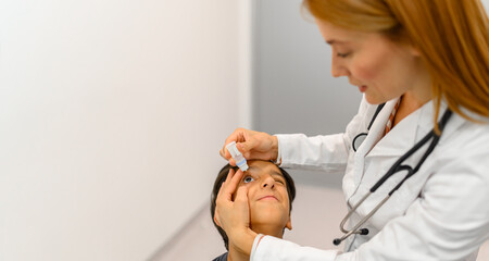 Female doctor putting eye drops in boy's eye during hospital visit at ophthalmology clinic for...
