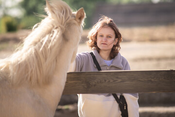 Young woman with wavy hair gently interacting with a white horse at a rustic fence, showcasing a serene connection between human and animal in a natural outdoor setting