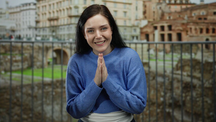 Woman with brunette hair meditating peacefully outdoors in front of beautiful historic roman ruins, wearing a blue sweater and smiling with hands in prayer position.