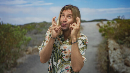 Obraz premium Blond man with long hair holds crossed fingers with both hands on a gravel path in a green park under blue sky; hope luck.
